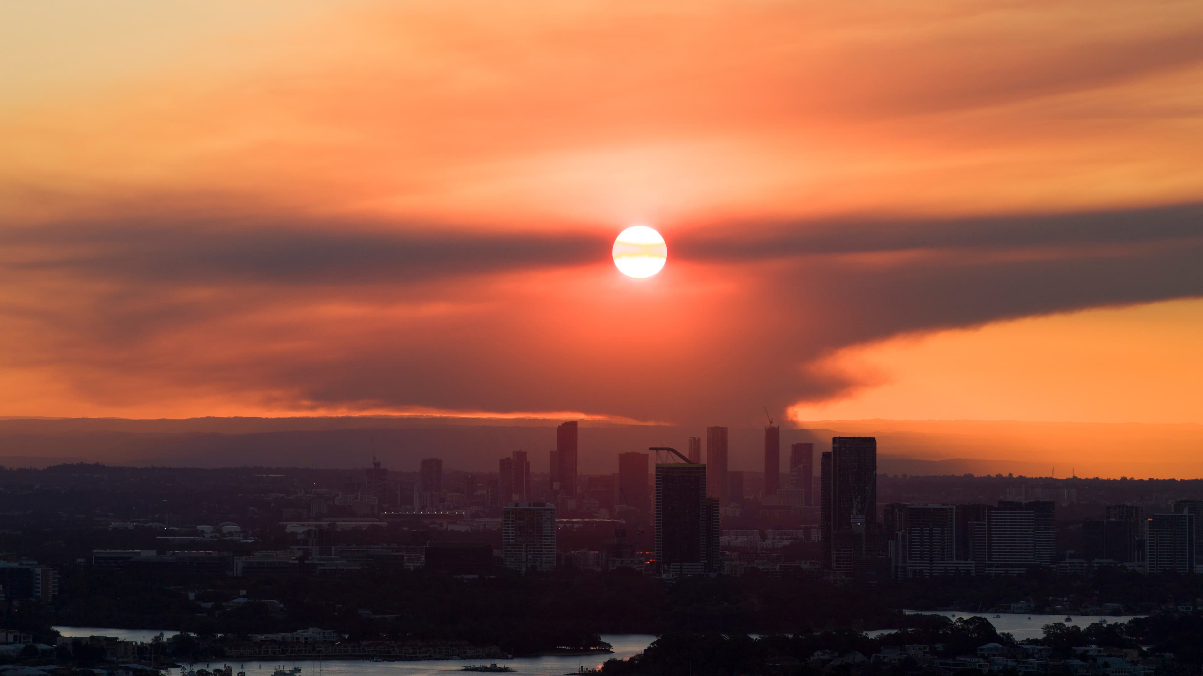 Bushfire smoke turning the sky orange and dimming the sun over an Australian city skyline, high-rise towers in silhouette