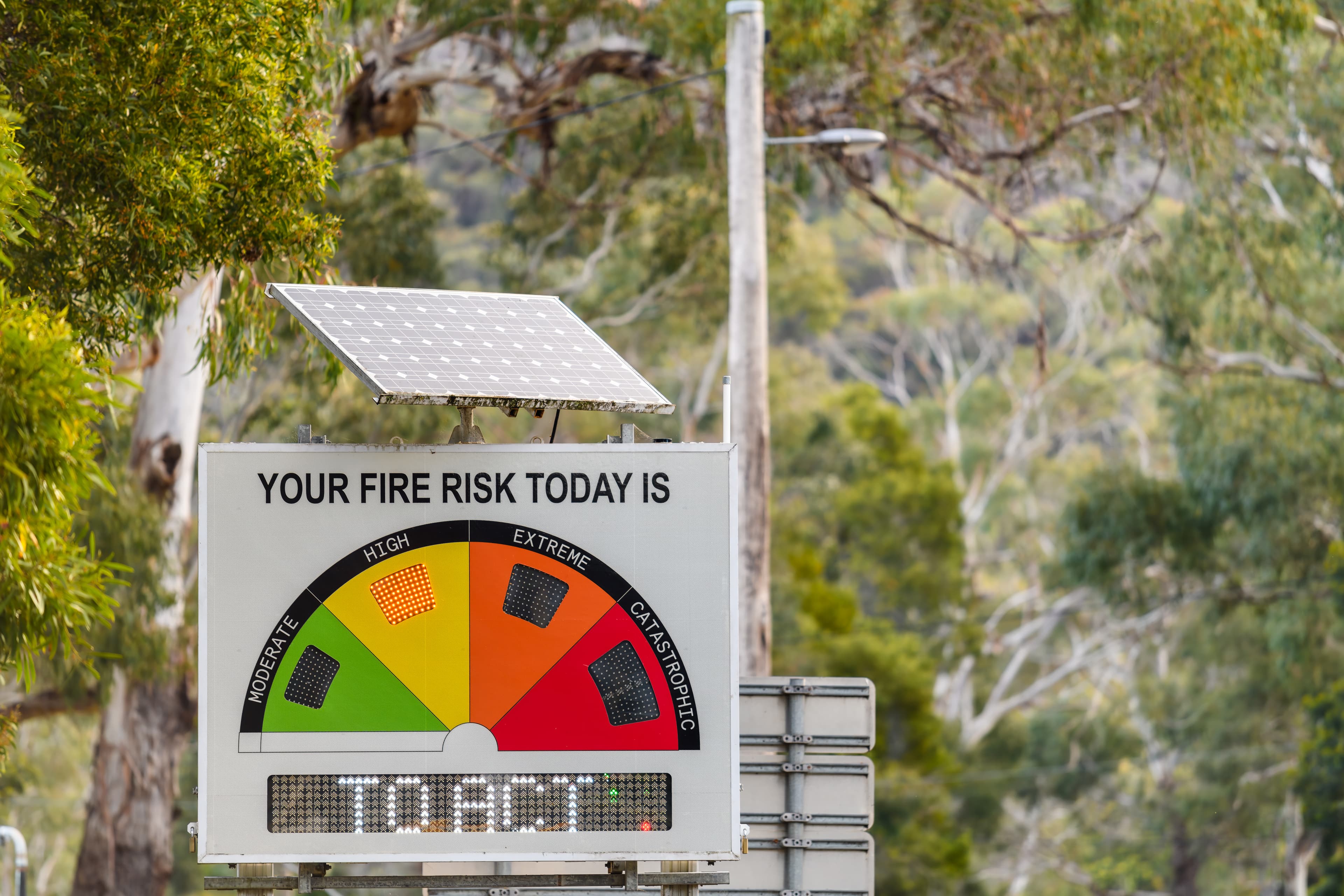 Solar-powered roadside Fire Danger Rating sign showing High on the Moderate to Catastrophic dial, with gum trees behind