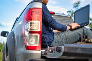 Person sitting on the open tailgate of a ute using a laptop outdoors against a rural backdrop.